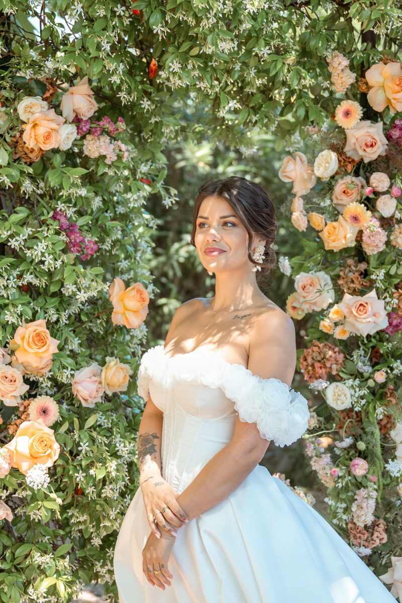 Jeune femme en robe de mariée devant une arche de cérémonie aux fleurs aux tons pêche Jeune femme en robe de mariée devant une arche de cérémonie fleurie - Raja Agency