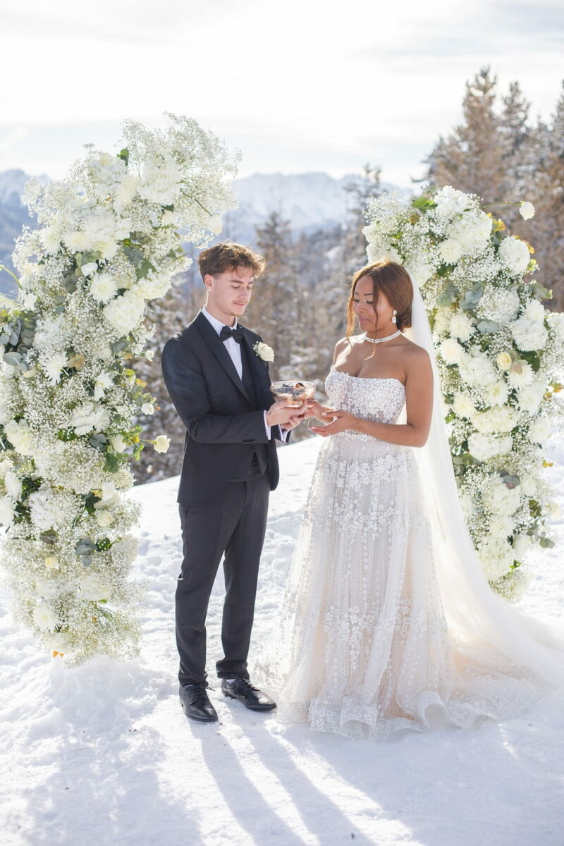 Couple de mariés devant leur arche de cérémonie en fleurs blanches, dans la neige Couple de mariés devant leur arche de cérémonie dans la neige - Raja Agency
