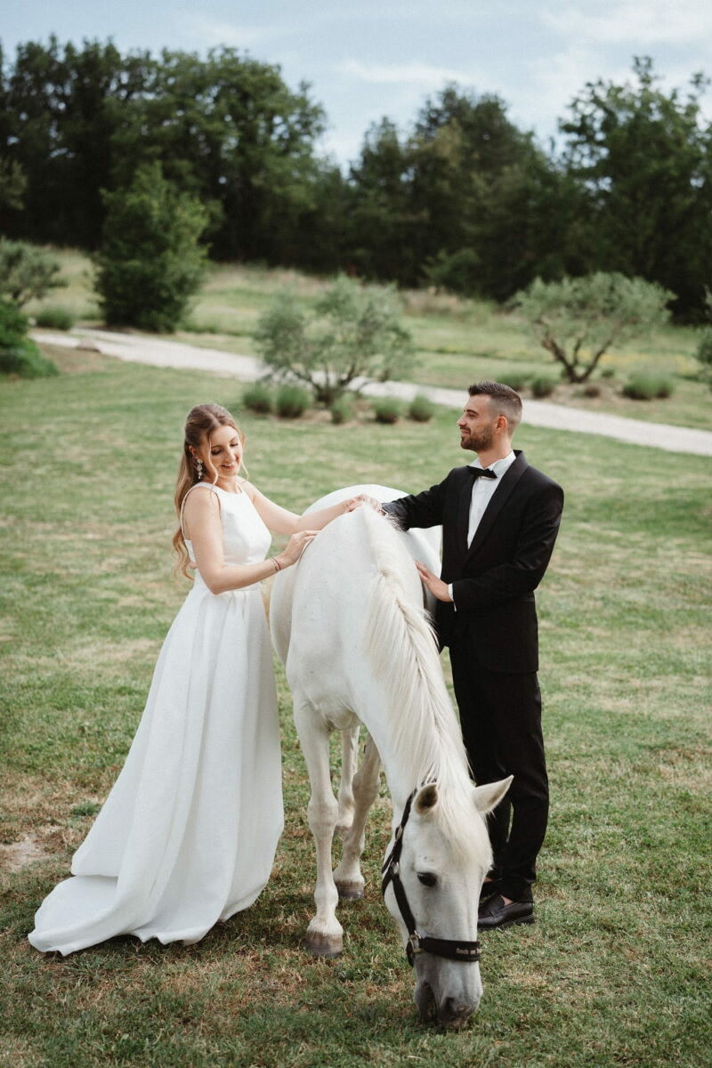 Couple de mariés souriant autour d'un cheval blanc, en extérieur Couple de mariés souriant autour d'un cheval - Raja Agency