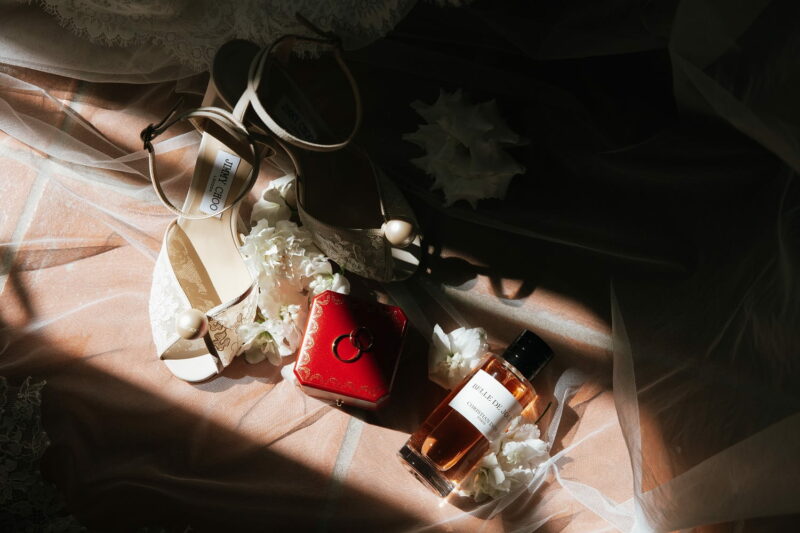 Flatlay de mariage contrasté, avec des talons Jimmy Choo et un écrin rouge, disposés sur un voile de mariée Flatlay de mariage contrasté, avec des talons Jimmy Choo et un écrin rouge - Raja Agency